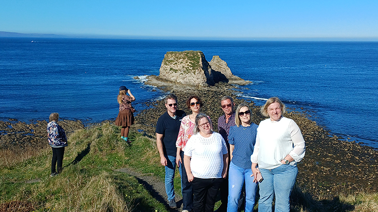 A group of people standing posing for a photo against a clifftop on a sunny day with the sea and sea stacks visible in the background