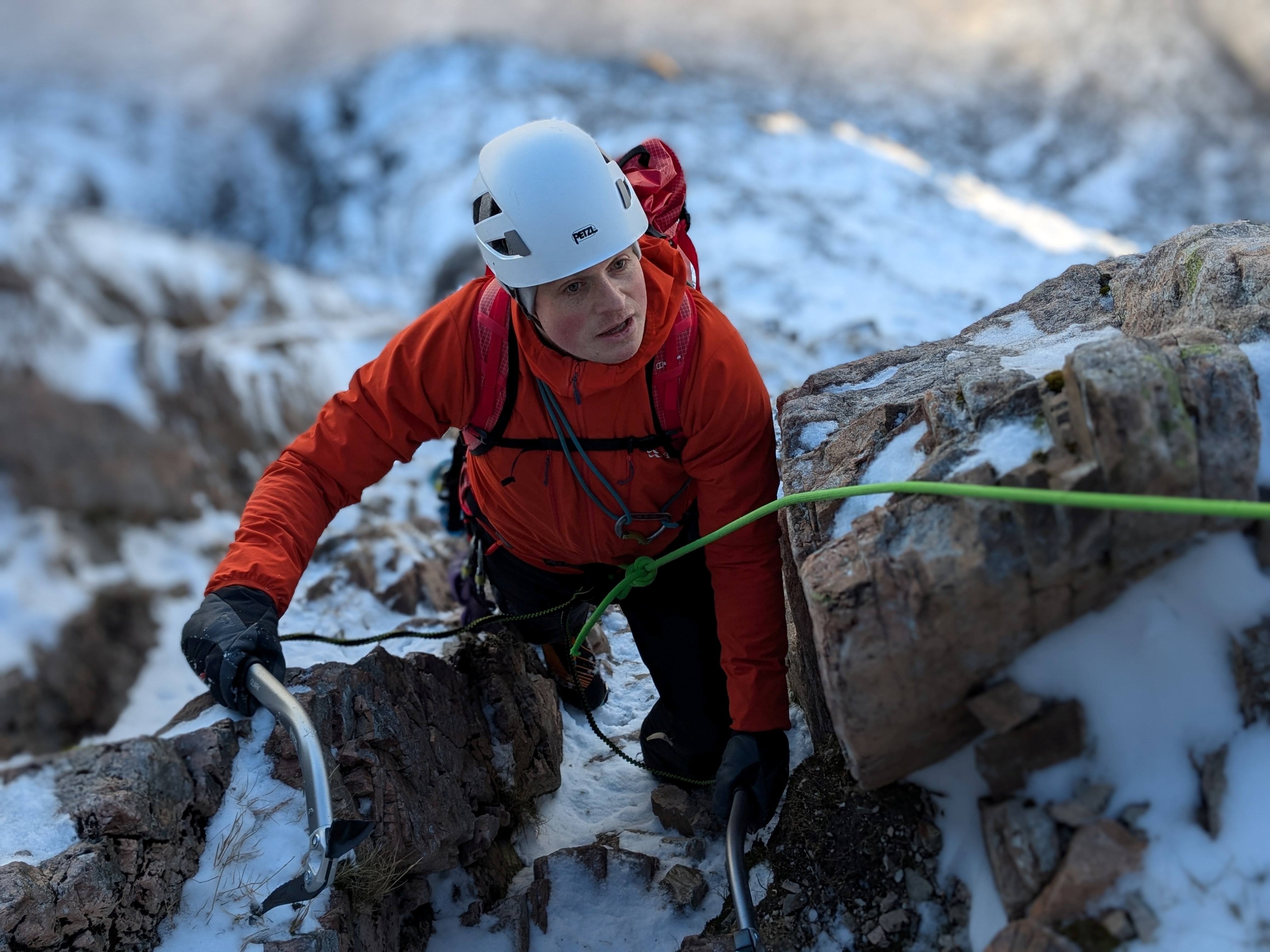 Person wearing a white helmet and orange jacket climbing a snowy mountain face using ice axes and ropes, with rocky terrain and a blurred winter landscape in the background.