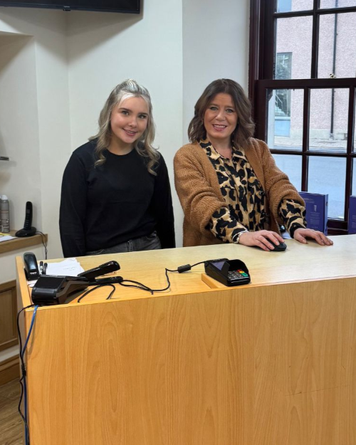 Apprentice Roni Wares, and employer Roslyn Hook, smiling behind a wooden service counter in Hair at 21, with a payment terminal and office supplies.