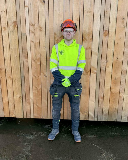 Modern apprentice Ryan Mackay standing in front of a timber-clad building, wearing full PPE including an orange safety helmet, high-visibility hoodie, and work gloves.