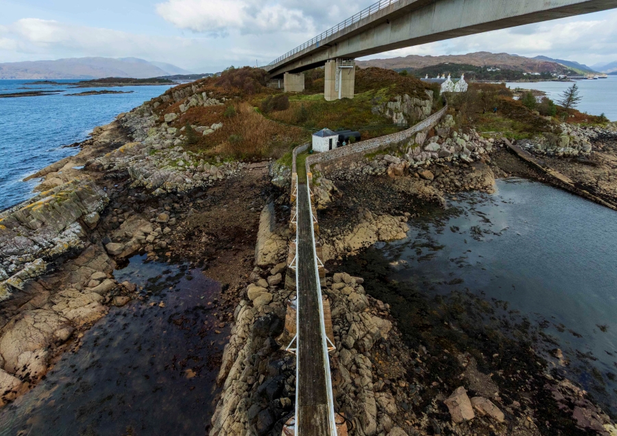 An overhead landscape photograph by photography student Brenna Young, showing a narrow wooden footbridge crossing a rocky coastline toward a small island, with a large concrete road bridge towering overhead.