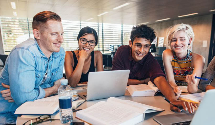 Mixed group of students sat round a table smiling and working on laptops